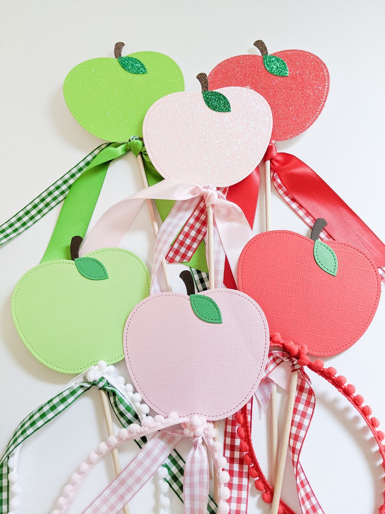 May include: Five apple-shaped paper crafts with glitter and ribbon accents. The apples are green, pink, and red. The ribbons are green and white gingham, pink and white gingham, and red and white gingham.