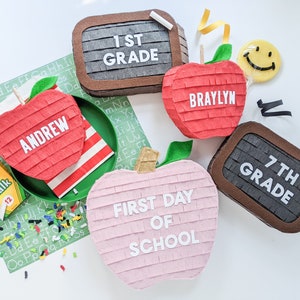 May include: A set of five colorful pinata decorations shaped like apples and a chalkboard. The pinatas are decorated with the words "1st Grade", "7th Grade", "First Day of School", "Braylyn", and "Andrew".