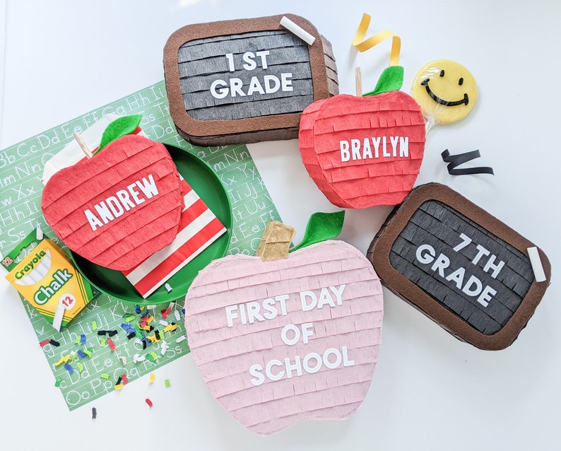 May include: A set of five colorful pinata decorations shaped like apples and a chalkboard. The pinatas are decorated with the words "1st Grade", "7th Grade", "First Day of School", "Braylyn", and "Andrew".