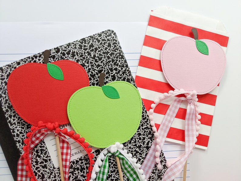 May include: Three paper apple cutouts on sticks with gingham ribbon and pom pom trim. The apples are red, green, and pink.