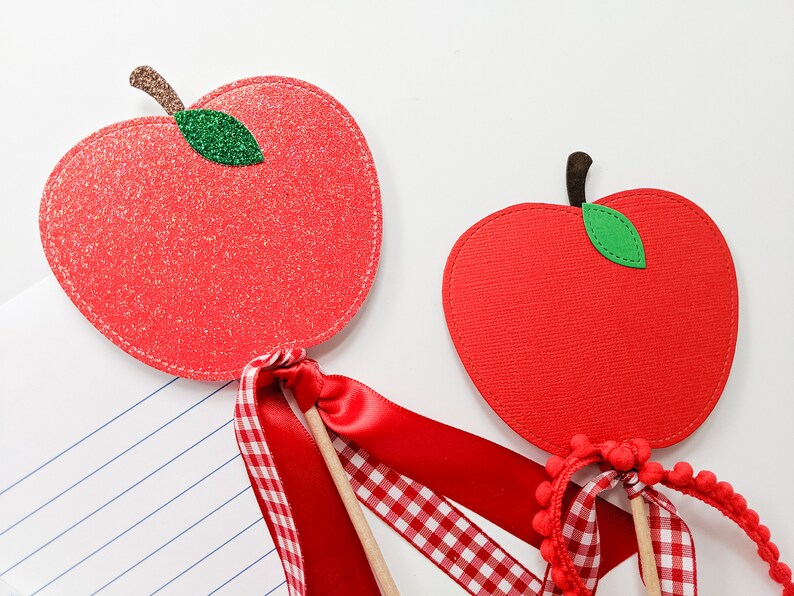 May include: Two red felt apple ornaments with green felt leaves. The apples are attached to wooden sticks with red and white gingham ribbon and red satin ribbon.