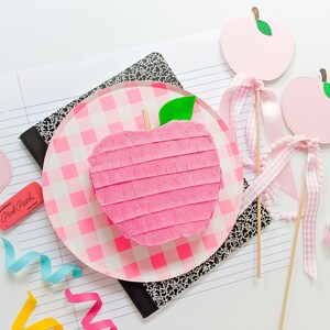 May include: Pink apple shaped pinata with a green leaf on top, sitting on a pink and white checkered plate. Two pink apple shaped decorations with green leaves and white gingham ribbon are on sticks.
