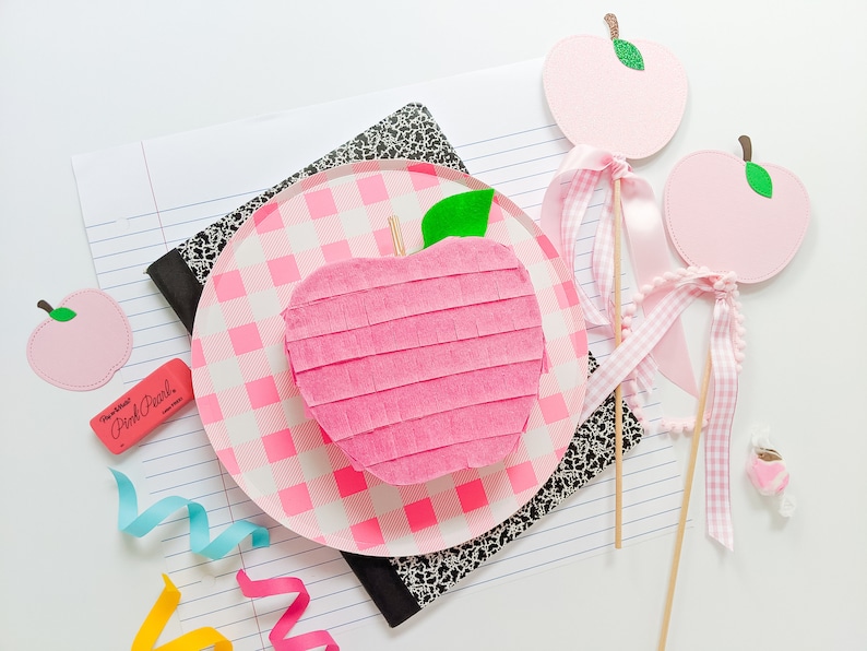May include: Pink apple shaped pinata with a green leaf on top, sitting on a pink and white checkered plate. Two pink apple shaped decorations with green leaves and white gingham ribbon are on sticks.