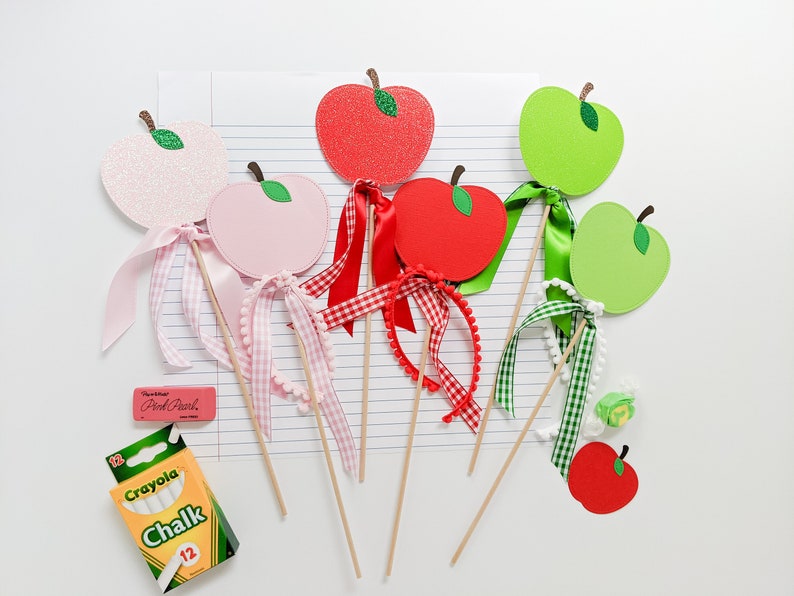 May include: Six apple-shaped craft sticks with glitter and ribbon embellishments. The apples are red, pink, and green. The ribbons are red and white gingham, pink, and green. There is a box of Crayola chalk and a pink eraser in the background.