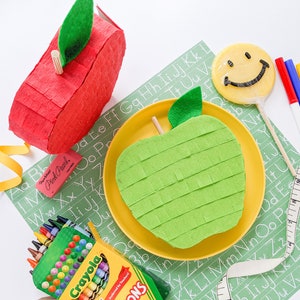 May include: Two apple-shaped pinatas, one red and one green, with a yellow plate and a smiley face lollipop. There are also crayons, markers, and a measuring tape on a green background with white letters.
