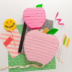 May include: Two pink apple-shaped pinatas with green leaves, a yellow smiley face lollipop, a pink and white striped paper bag with a red eraser that says "Proku", a black and white notebook, a green sheet of paper with the alphabet printed on it, a pink ribbon, and three highlighters in green, yellow, and white.