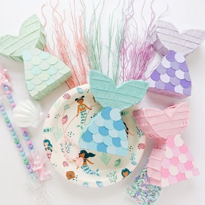May include: Five mermaid tail shaped pinatas in various shades of pink, blue, and green. The pinatas are decorated with paper scales and fringe. There are also mermaid themed party supplies, including a paper plate with mermaids, a shell, and a beaded wand.