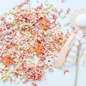 May include: A pink and white flower wand with a white pom-pom on top. The wand is surrounded by confetti in shades of pink, yellow, and orange. The confetti includes peace signs, flowers, and the words "Groovy" and "Peace".