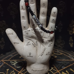 May include: A white ceramic hand with lines and symbols representing different aspects of palmistry. The hand is wearing a black beaded bracelet with red and silver accents. The hand is sitting on a black and gold circular surface with symbols.