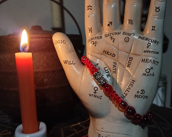 May include: A white ceramic hand with lines and symbols representing the lines of a palm reading. The hand is wearing a red beaded bracelet with a silver charm. The hand is sitting on a black surface with a white candle holder with a pentagram symbol.