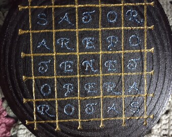 Puede incluir: Un círculo de madera con una cuadrícula dorada y letras bordadas azules que deletrean la frase latina "SATOR AREPO TENET OPERA ROTAS".