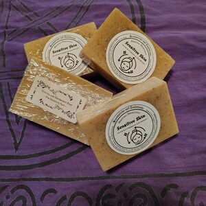 May include: Three rectangular bars of tan, speckled soap wrapped in clear plastic. Each bar features a circular white label with the text Broomsticks and Toadstools, Magical Medicinals, Sensitive Skin, with buttermilk and honey.