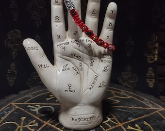 May include: A white ceramic hand with lines and symbols representing different aspects of palmistry. The hand is wearing a red and silver bracelet with beads. The hand is sitting on a black surface with a gold and red design.