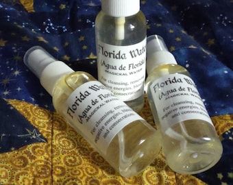 Pode incluir: Três garrafas de vidro transparente com tampas douradas e etiquetas brancas. As etiquetas têm texto preto que diz "Florida Water (Agua de Florida) MAGICKAL WATER" e instruções de uso. As garrafas estão sobre um fundo de tecido azul e amarelo.