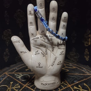 Puede incluir: Una mano de cerámica blanca con símbolos y marcas astrológicas, sosteniendo un brazalete de cuentas azules y negras. La mano está sentada sobre una superficie negra con símbolos dorados y rojos.