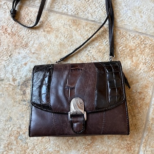 May include: A brown leather crossbody bag with a long strap. The bag features a mix of textures, including smooth and embossed leather, with a silver-tone clasp. The bag is approximately 8 inches wide and 6 inches tall.