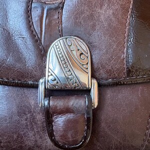 May include: Close-up of a brown leather handbag with a decorative silver clasp. The clasp features an ornate design with etched details. The leather has a textured appearance with visible stitching and a glossy finish.