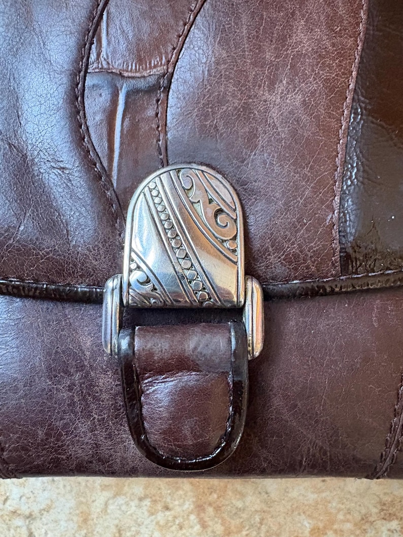 May include: Close-up of a brown leather handbag with a decorative silver clasp. The clasp features an ornate design with etched details. The leather has a textured appearance with visible stitching and a glossy finish.