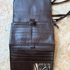 May include: Open brown leather crossbody wallet with multiple card slots and a clear ID window. The wallet has a flap closure with a decorative button and a long, braided strap. The interior is lined with a lighter brown material.