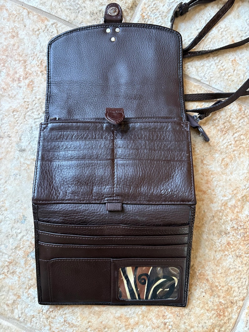May include: Open brown leather crossbody wallet with multiple card slots and a clear ID window. The wallet has a flap closure with a decorative button and a long, braided strap. The interior is lined with a lighter brown material.