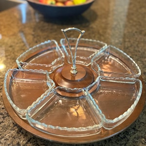 May include: A circular wooden serving tray featuring five removable, wedge-shaped clear glass dishes with scalloped edges. A central metal handle with a silver-toned finish is attached to the wooden base, allowing for easy transport of the serving set.