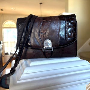 May include: A brown leather crossbody bag with a textured flap and a silver-tone clasp. The bag features a long, adjustable strap and is displayed on a white surface. The bag's design includes a mix of smooth and embossed leather.