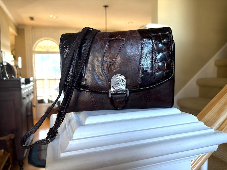 May include: A brown leather crossbody bag with a textured flap and a silver-tone clasp. The bag features a long, adjustable strap and is displayed on a white surface. The bag's design includes a mix of smooth and embossed leather.