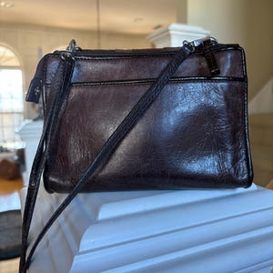 May include: A dark brown leather crossbody bag with a textured finish. The bag has a zippered top closure, a front pocket, and a long, adjustable strap. The hardware is silver-toned.