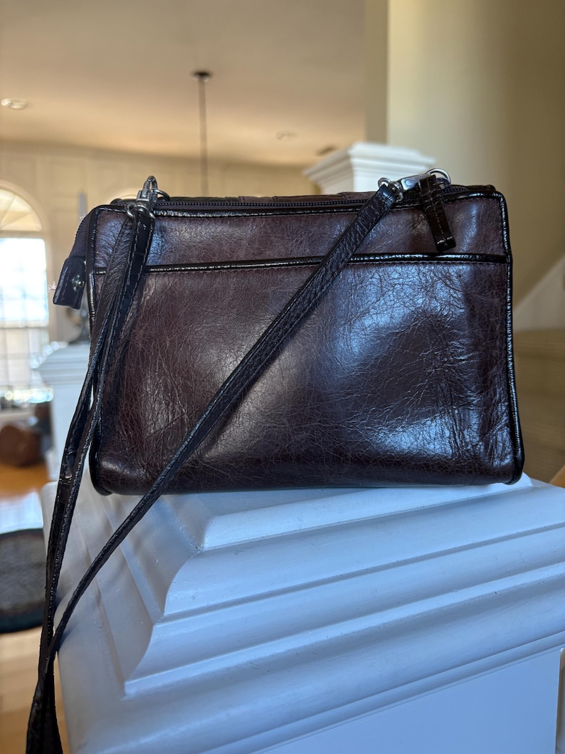 May include: A dark brown leather crossbody bag with a textured finish. The bag has a zippered top closure, a front pocket, and a long, adjustable strap. The hardware is silver-toned.