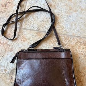 May include: A brown leather crossbody bag with a long, adjustable strap. The bag has a rectangular shape with a zippered top and a front pocket. The leather has a textured appearance. The strap is thin and dark brown.