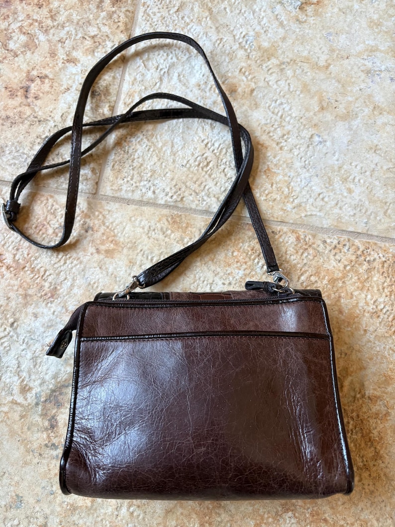 May include: A brown leather crossbody bag with a long, adjustable strap. The bag has a rectangular shape with a zippered top and a front pocket. The leather has a textured appearance. The strap is thin and dark brown.
