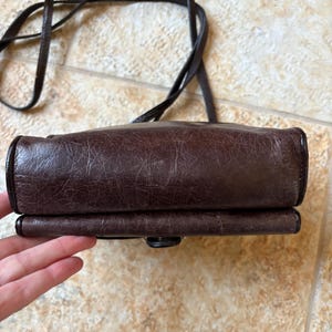 May include: A dark brown leather crossbody bag with a long strap. The bag has a rectangular shape with rounded edges and a textured surface. The strap is thin and made of the same leather material.