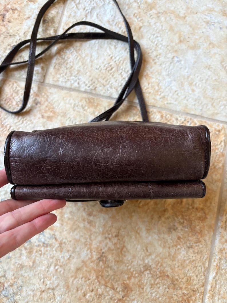 May include: A dark brown leather crossbody bag with a long strap. The bag has a rectangular shape with rounded edges and a textured surface. The strap is thin and made of the same leather material.