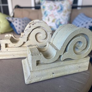 May include: Two decorative, white, wooden corbels with a scroll design. The corbels are painted a light cream color and have a distressed finish.
