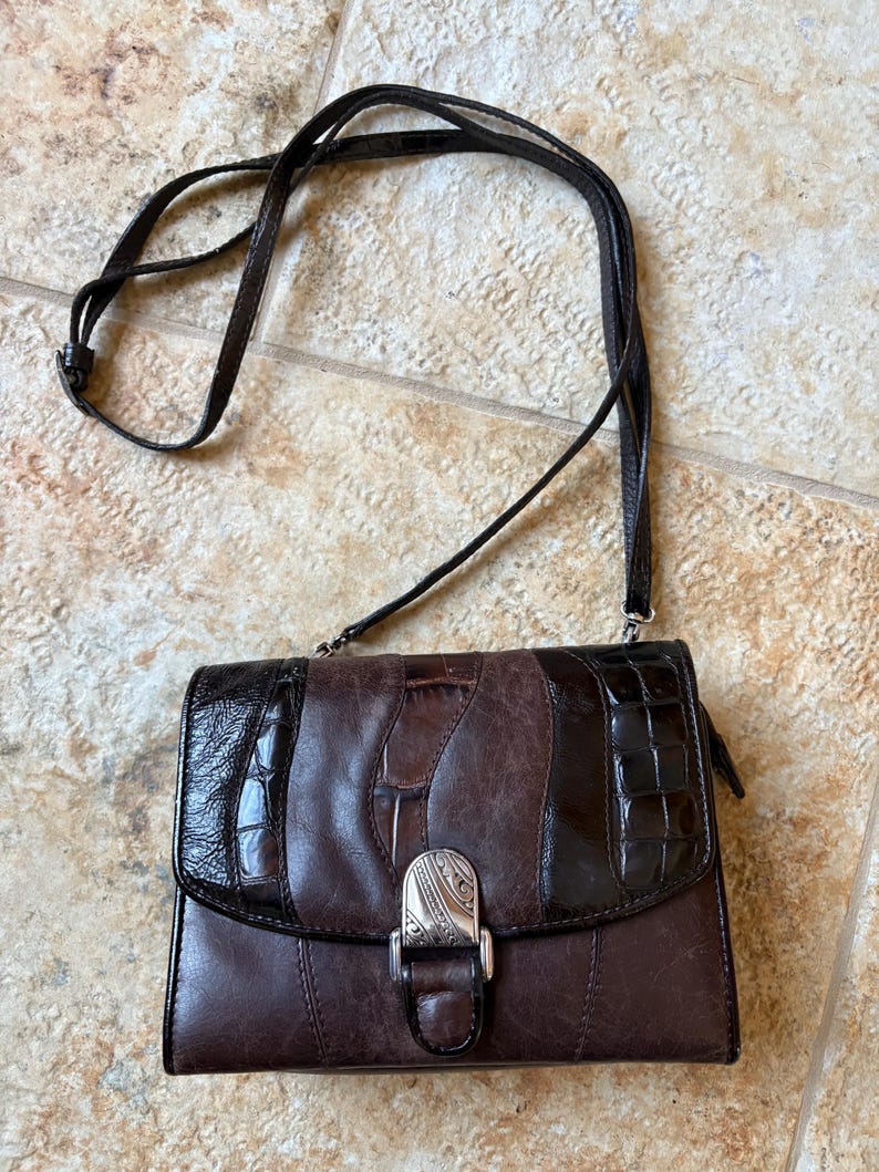 May include: A brown leather crossbody bag with a long strap. The bag features a mix of textures, including smooth and embossed leather, with a silver-tone clasp. The bag is approximately 8 inches wide and 6 inches tall.