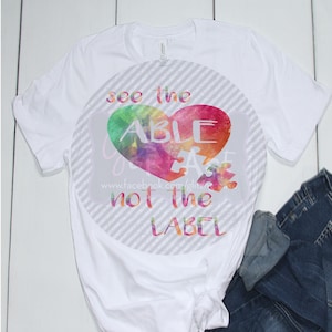 May include: White t-shirt with a round design featuring a watercolor heart with the word "ABLE" inside and a puzzle piece. The design also includes the text "see the" above the heart and "not the LABEL" below the heart.