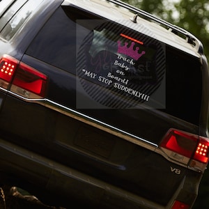Peut inclure: Un SUV Toyota noir avec un autocollant "Trash Baby on Board! May Stop Suddenly!!!" sur la vitre arrière. L'autocollant présente une couronne rose pailletée et du texte en écriture cursive.