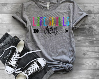 Puede incluir: Camiseta gris con el texto "Life Skills Crew" en letras de colores con una flecha negra apuntando hacia la derecha. La camiseta está sobre una superficie de madera con un par de zapatillas negras y unos vaqueros azules.