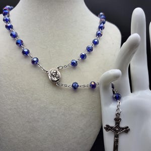 May include: A rosary necklace with faceted, deep blue beads and a silver-toned crucifix pendant. The necklace is displayed on a white mannequin hand and a bust. The cross features a detailed depiction of Jesus.