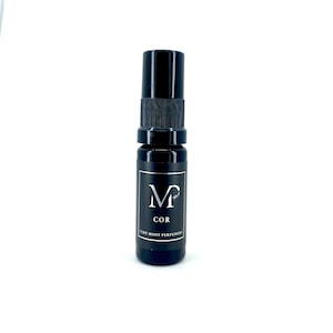 May include: A black glass bottle with a black spray nozzle and a white label. The label has the text "M" with a small design above it, "COR" below it, and "VERT MONT PERFUMERY" at the bottom.