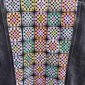 May include: A black denim jacket with a colorful, patchwork design on the back. The design features a grid of crochet squares in pastel colors including pink, yellow, orange, and green. The jacket has a classic collar and button closure.
