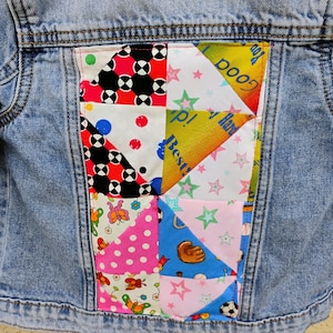 May include: A light blue denim jacket with a patchwork design on the back. The patchwork features various colourful patterns, including polka dots, stars, and geometric shapes. The text "Good" and "Best" are visible.