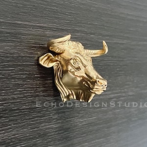 May include: A gold-colored bull head sculpture with detailed features. The sculpture is mounted on a dark wood surface.