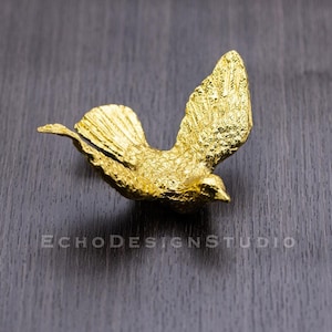 Puede incluir: Una figurita de pájaro de color dorado con una superficie texturizada. El pájaro está en vuelo con las alas extendidas.