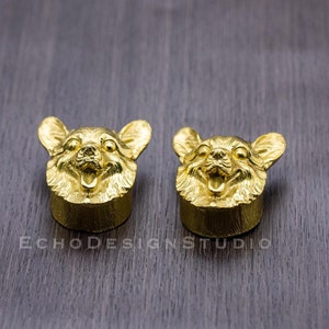 May include: Two gold-colored, detailed, three-dimensional dog head bottle stoppers. The dog has a short, fluffy coat and is smiling with its tongue sticking out.