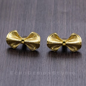 May include: A pair of gold bow-shaped earrings. The earrings are made of a textured metal and have a shiny finish.
