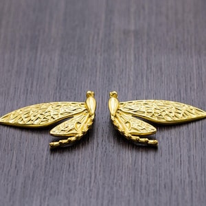 Può includere: Un paio di orecchini a forma di libellula dorati con dettagli intricati. Gli orecchini sono su uno sfondo di legno scuro.