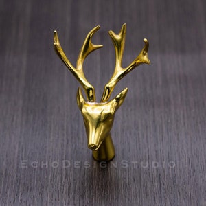 May include: A gold-coloured deer head figurine with detailed antlers. The deer head is centered against a dark wood-grain background. The text "ECHODESIGNSTUDIO" is visible at the bottom.
