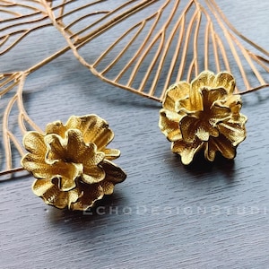 May include: Two gold-colored flower-shaped decorative objects on a dark gray wooden surface. The objects are made of metal and have a textured surface.