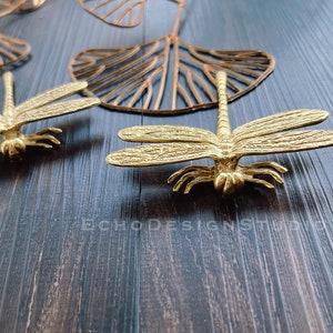 Könnte beinhalten: Zwei goldene Libellen aus Metall als Wanddekoration auf einem dunklen Holzgrund. Die Libellen haben detaillierte Flügel und Beine.